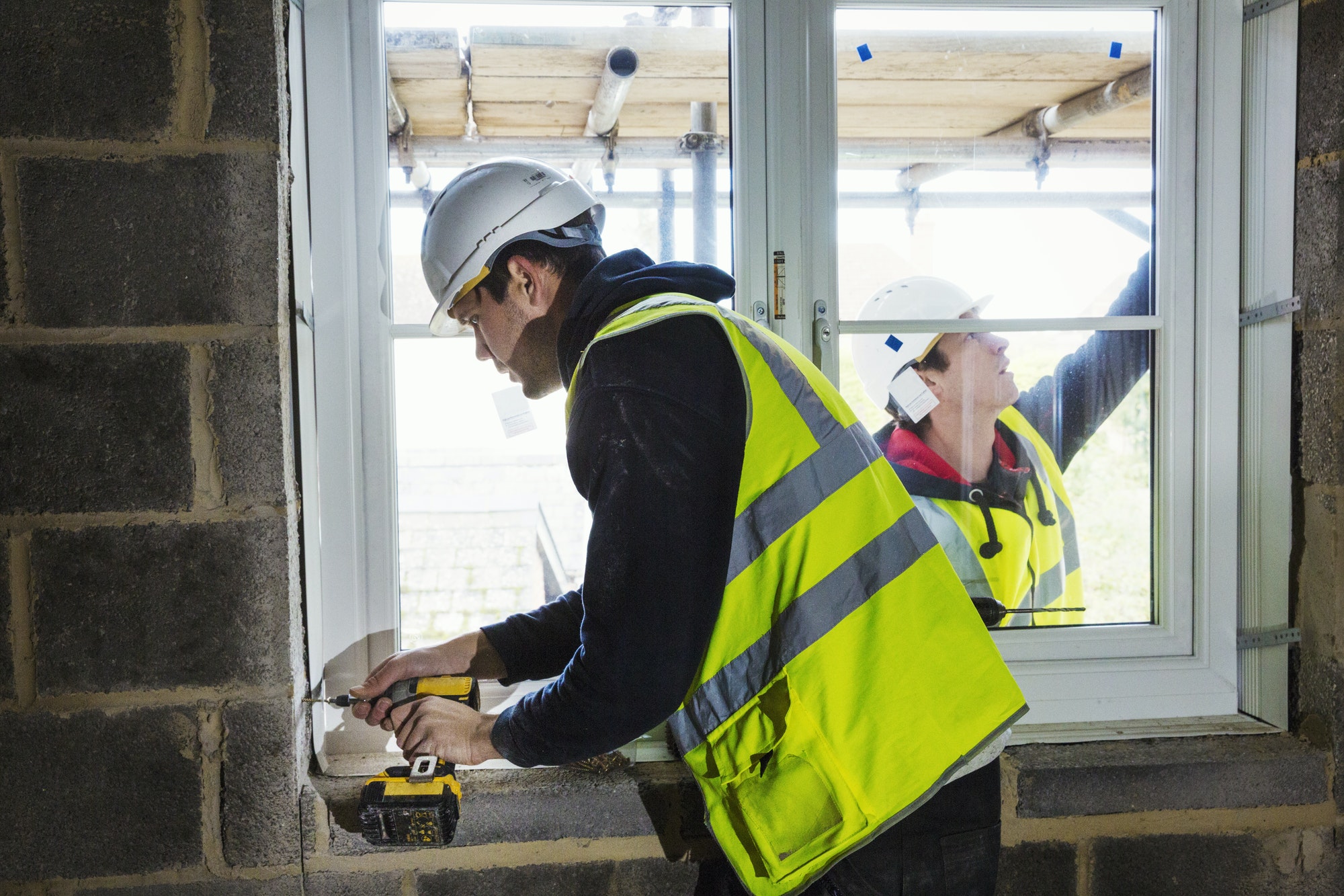 A workman on a construction site, builder in hard hat using an electric drill on a window sill.