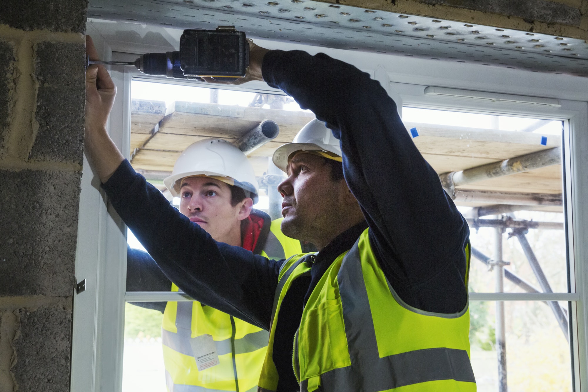 Two workmen on a construction site, builder in hard hat using an electric drill on a window frame.